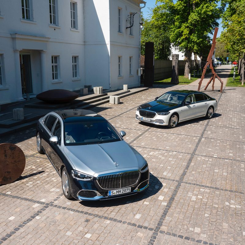 Two 2022 Mercedes-Maybach S-Class sedans parked in front of a white mansion on a brick parking pad with sculptures in the back.