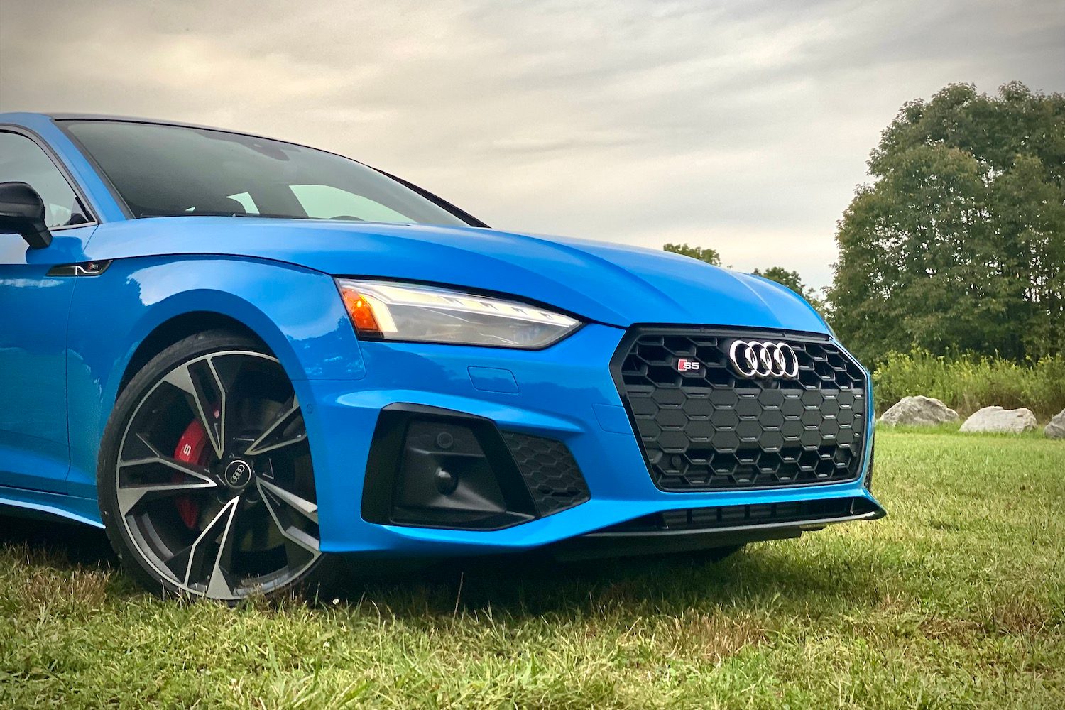 Front end angle of 2023 Audi S5 Sportback from passenger's side in a grassy field during sunset.