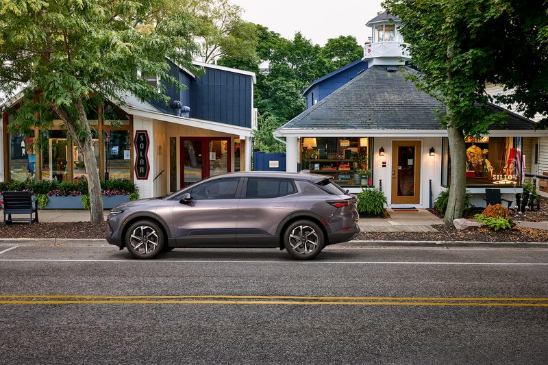 Side profile of the 2024 Chevrolet Equinox EV 1LT in front of buildings while driving down the road.