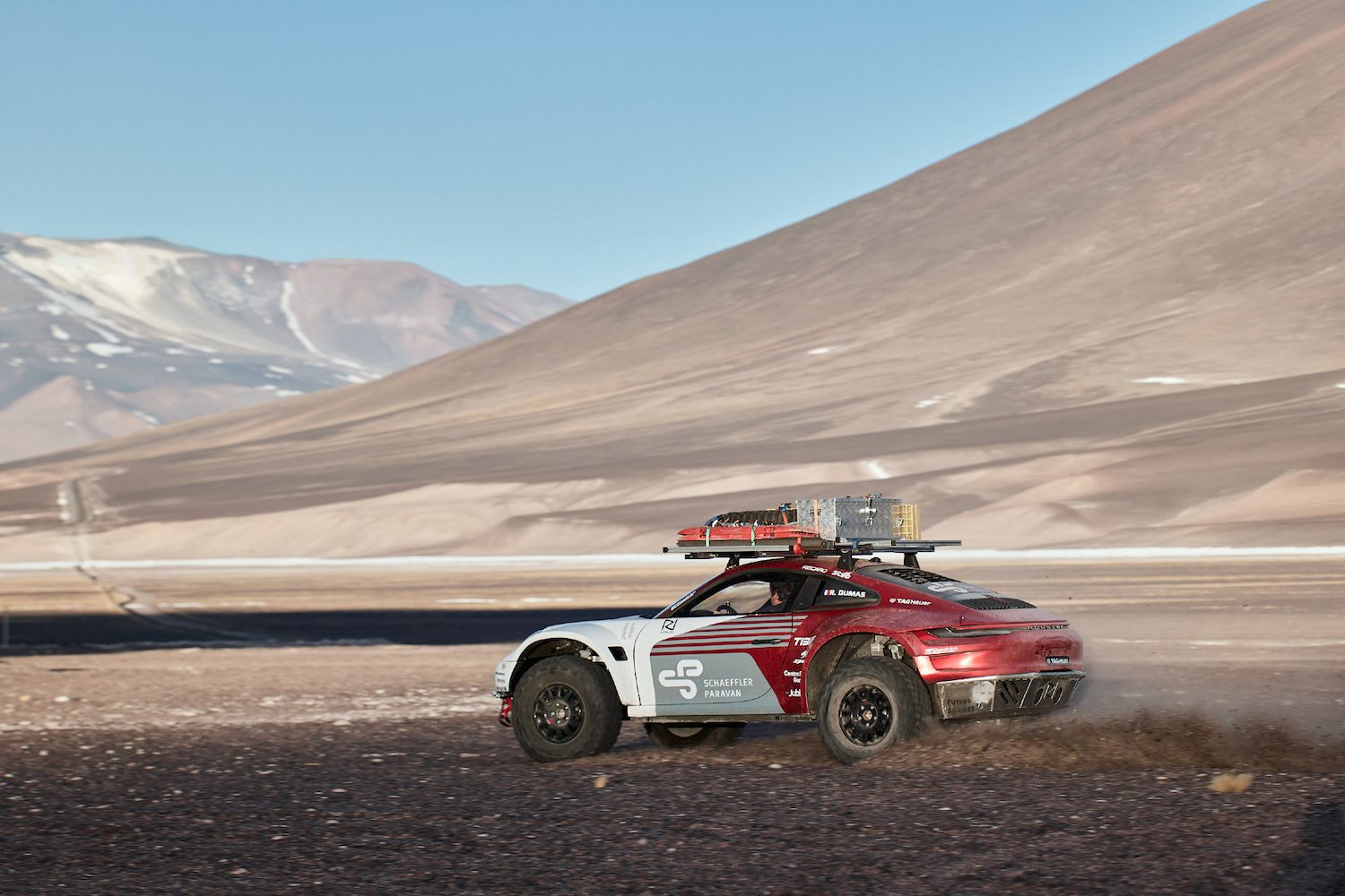 Rear end angle of Porsche 911 Dakar Prototype traveling quickly along rocky terrain with mountains in the back.