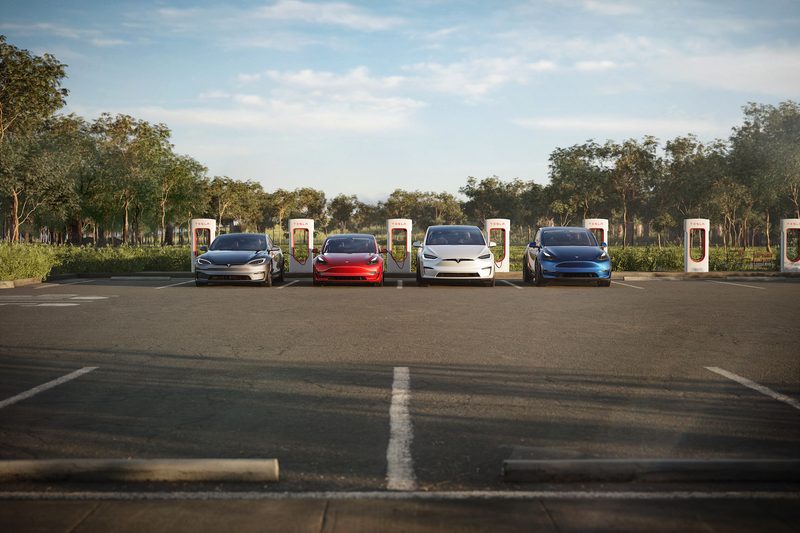 Tesla's lineup of electric vehicles lined up in front of chargers in a parking lot with trees in the back.