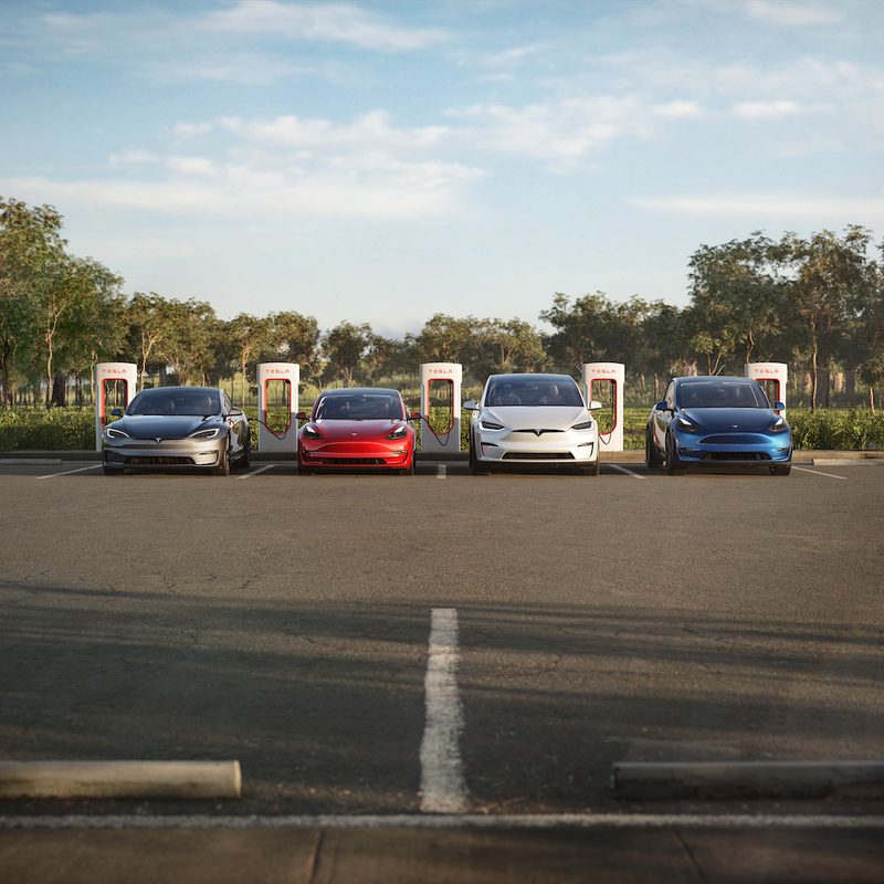 Tesla's lineup of electric vehicles lined up in front of chargers in a parking lot with trees in the back.