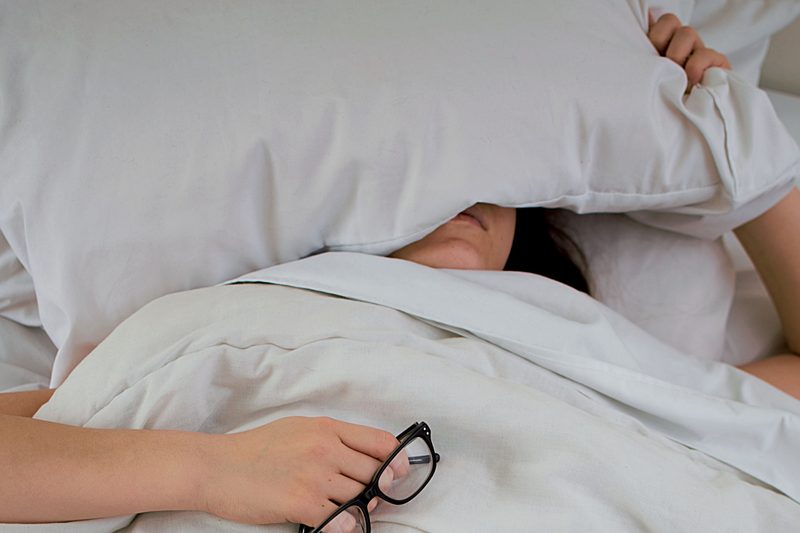 person sleeping with head under pillow and glasses in their hand