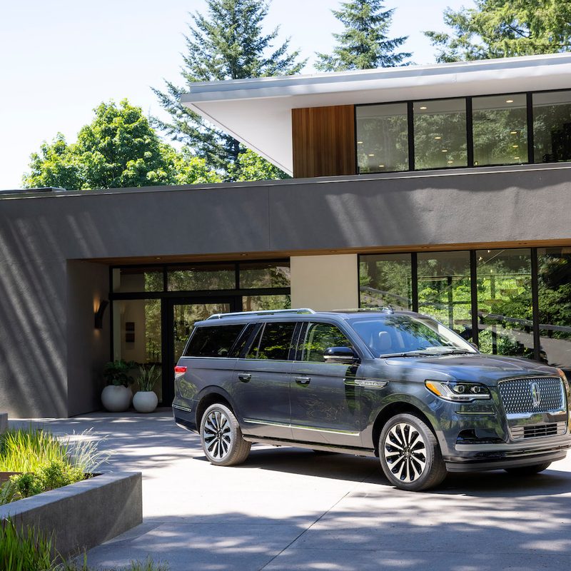 2022 Lincoln Navigator Flight Blue Reserve front end angle from passenger's side in front of a mansion with trees and blue skies in the back.