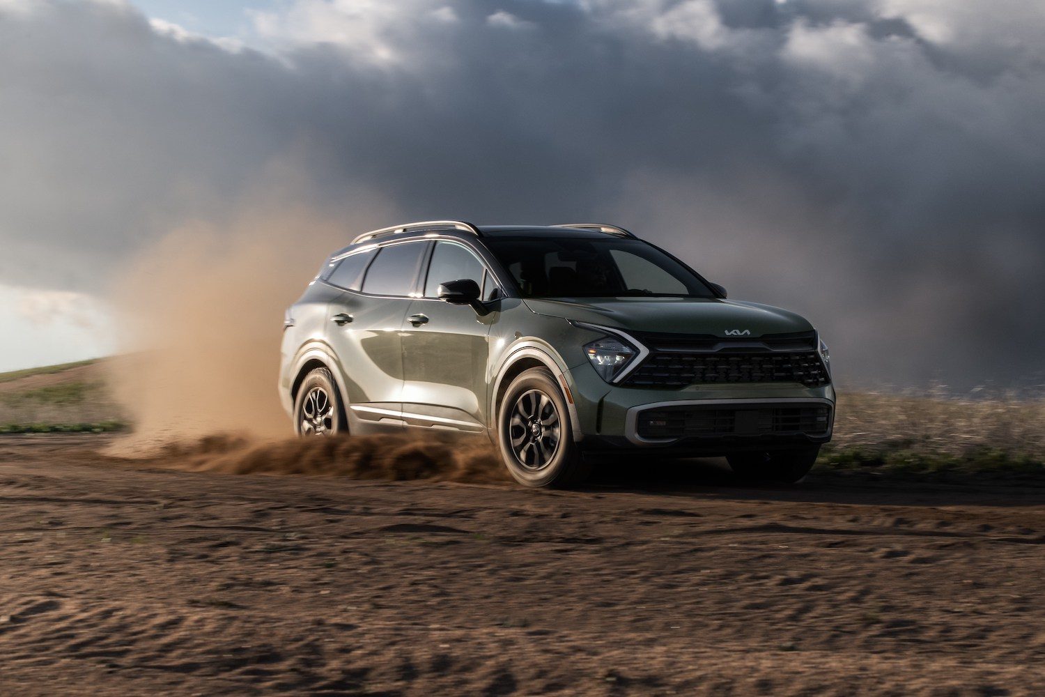 2023 Sportage X-Pro front end angle from passenger side driving on a dirt road with dirt flying up in the back and dark clouds in the sky.
