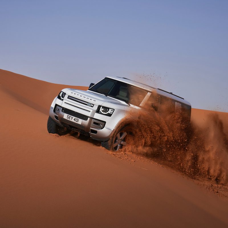 2023 Land Rover Defender 130 front end driving across a sand dune in the desert with blue skies in the back.