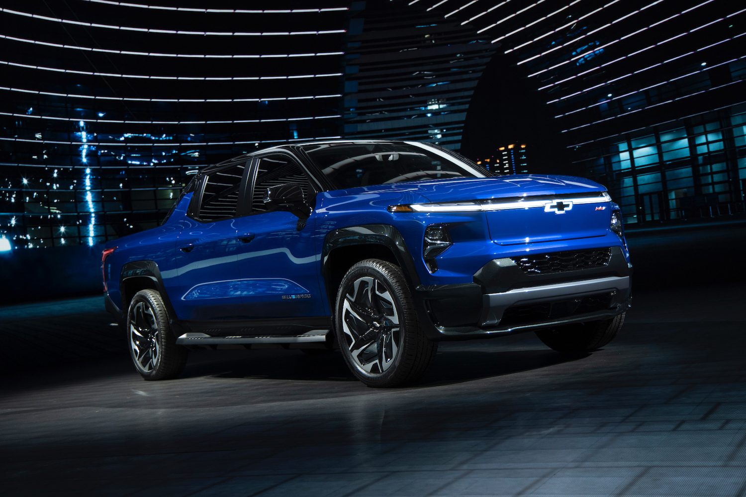 2024 Chevrolet Silverado EV RST pickup truck front end angle from passenger's side parked under a light with large window and buildings in the back.