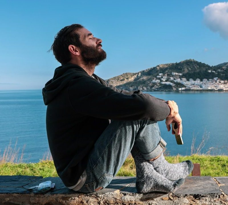 Sad man sitting on the ground near a body of water with his eyes closed.