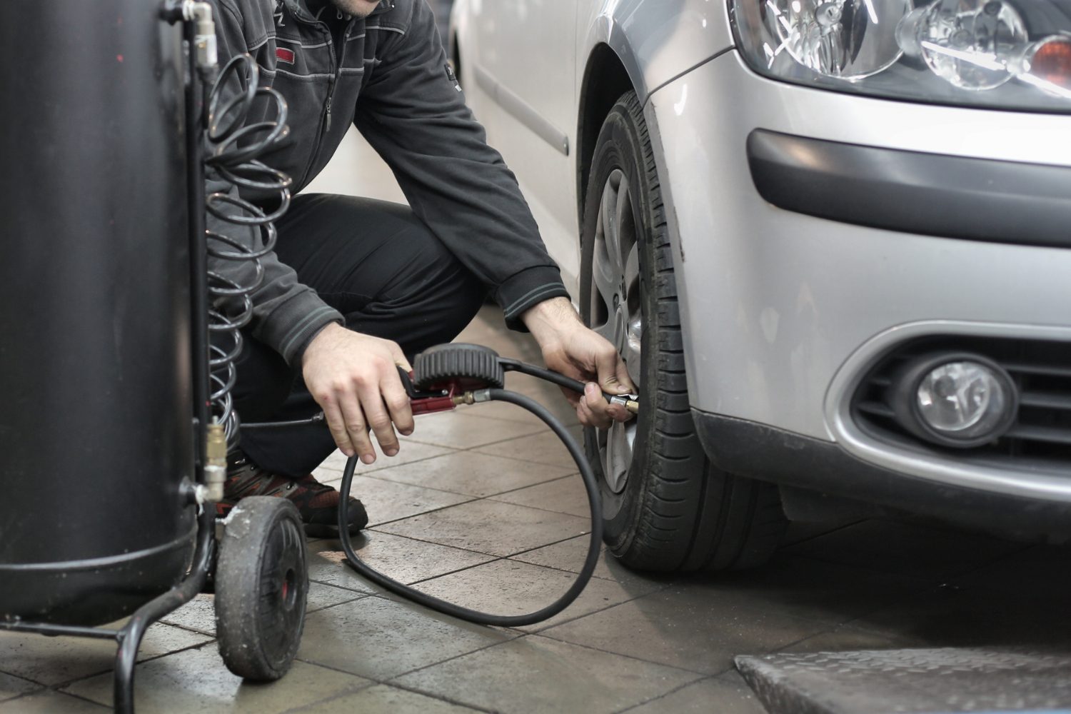 Man in black checking tire pressure on tires with a car parked in a garage.