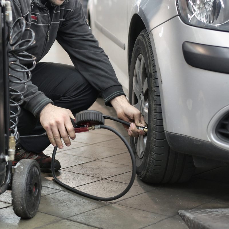 Man in black checking tire pressure on tires with a car parked in a garage.
