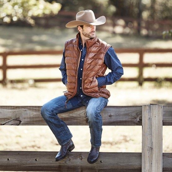A man sitting on a fence post and wearing jeans, a polka-dot dress shirt, a brown vest, an da tan cowboy hat