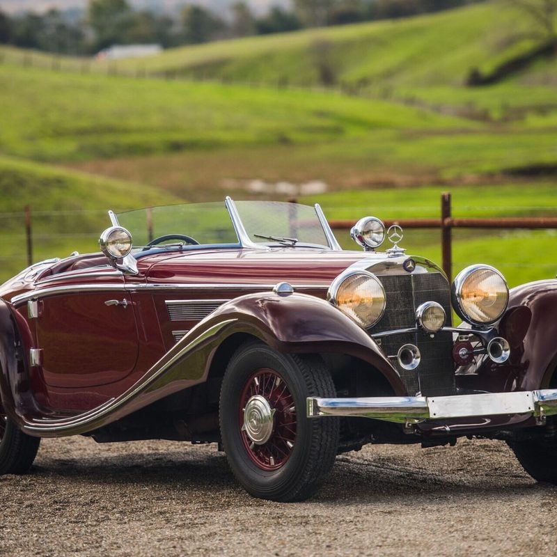 1937 Mercedes-Benz 540 K Special Roadster front end angle from passenger side parked on a dirt path with green fields in the back.