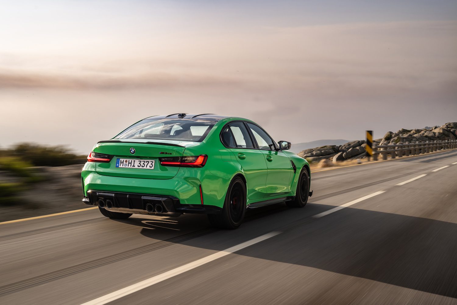 Rear end angle of 2024 BMW M3 CS driving down a road with clouds in the back.