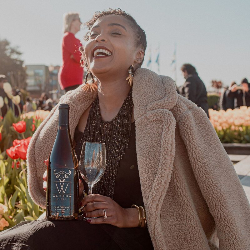 A Black woman wearing a black dress and tan coat holding a bottle of wine and a wine glass