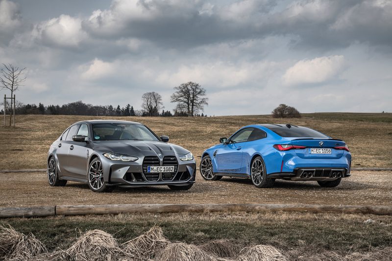 2022 BMW M3 Competition xDrive and M4 Competition xDrive parked in a dirt field in front of dirt patches with dark clouds in the back.