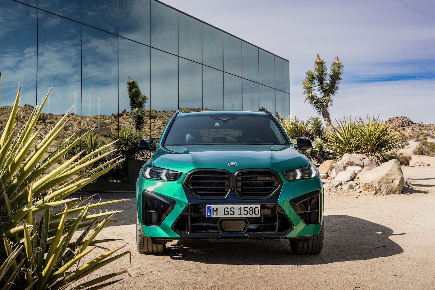 2024 BMW X5 M Competition front end close up parked in front of a glass building with desert in the back.