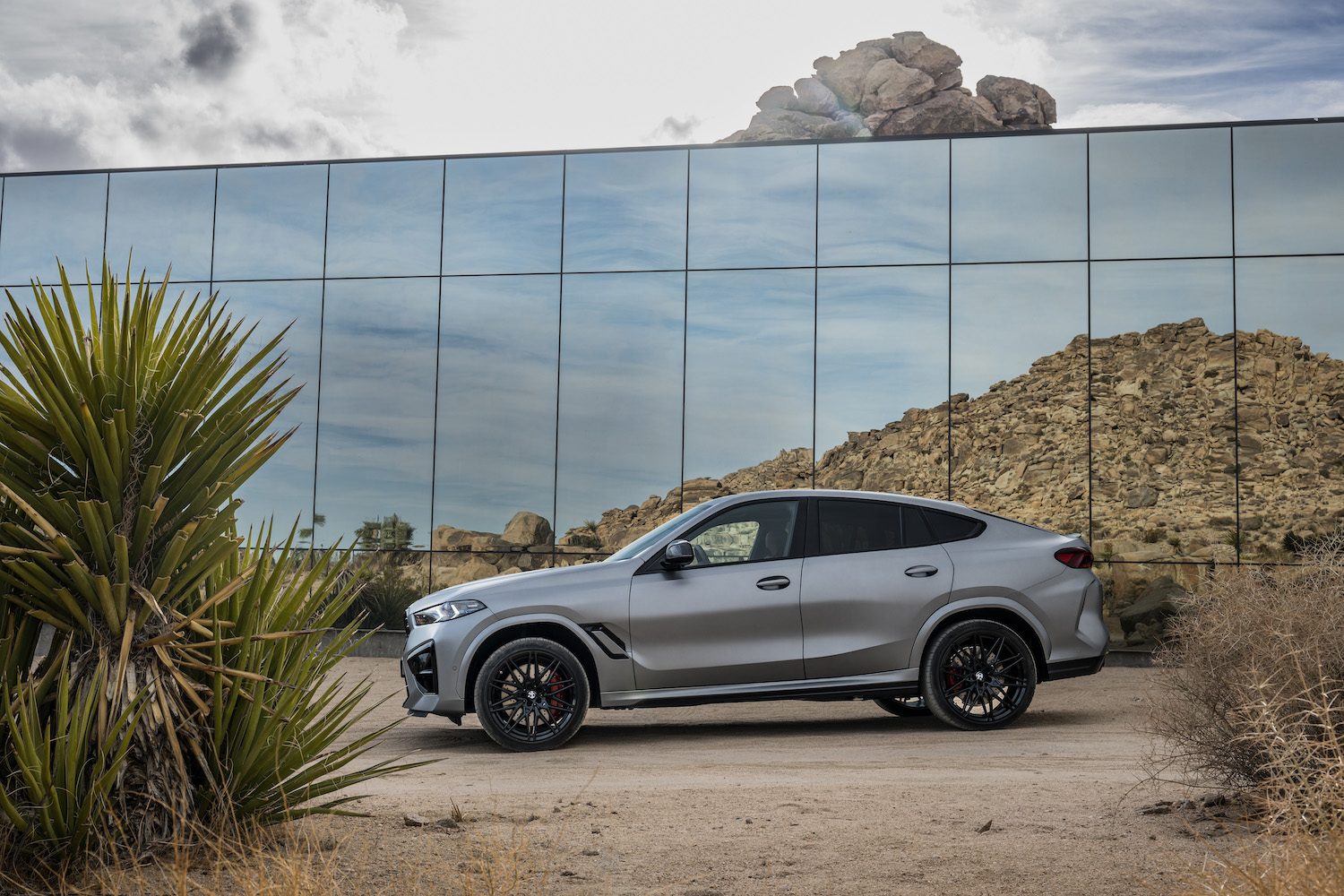 Side profile of the 2024 BMW X6 M Competition parked in the desert in front of a glass building.
