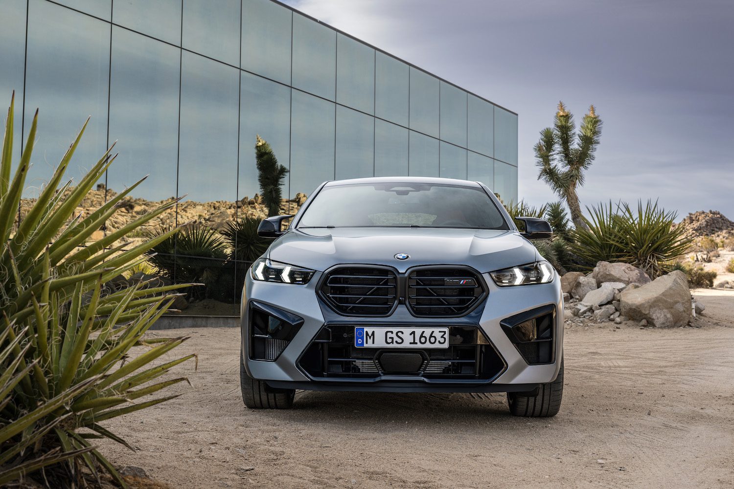 2024 BMW X6 M Competition front end close up parked in front of a glass building in the desert.