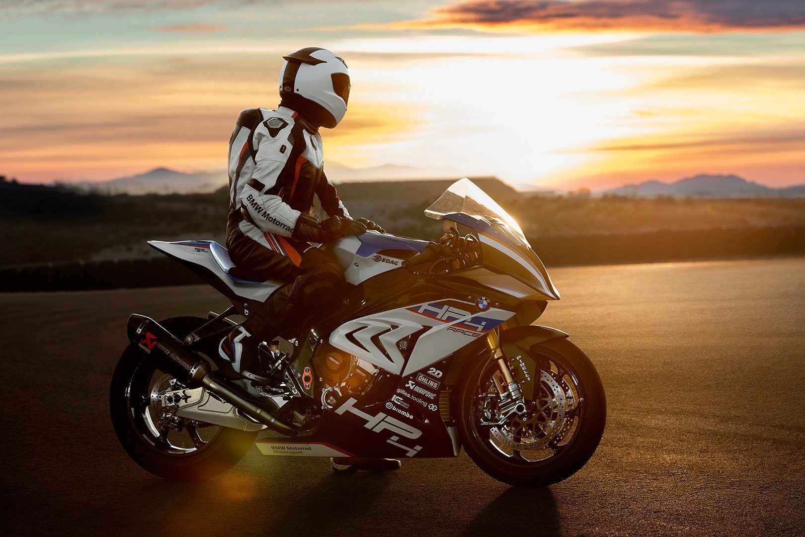 BMW HP4 Race super bike with a rider sitting on the motorcycle looking at a sunset at the track.