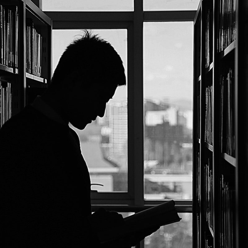 A man in silhouette reads a book between two large bookshelves in front of a window.