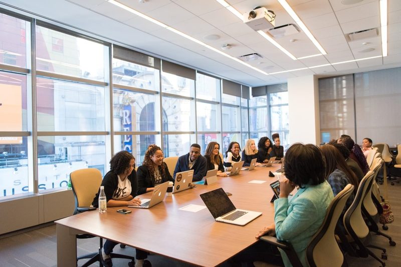 A big group all sitting at a large desk with people on their laptops in a large office room.