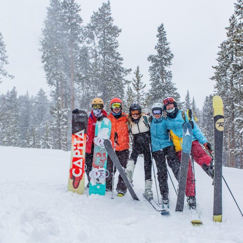 A group of friends posing with their snowboards and skis while standing in the snow.