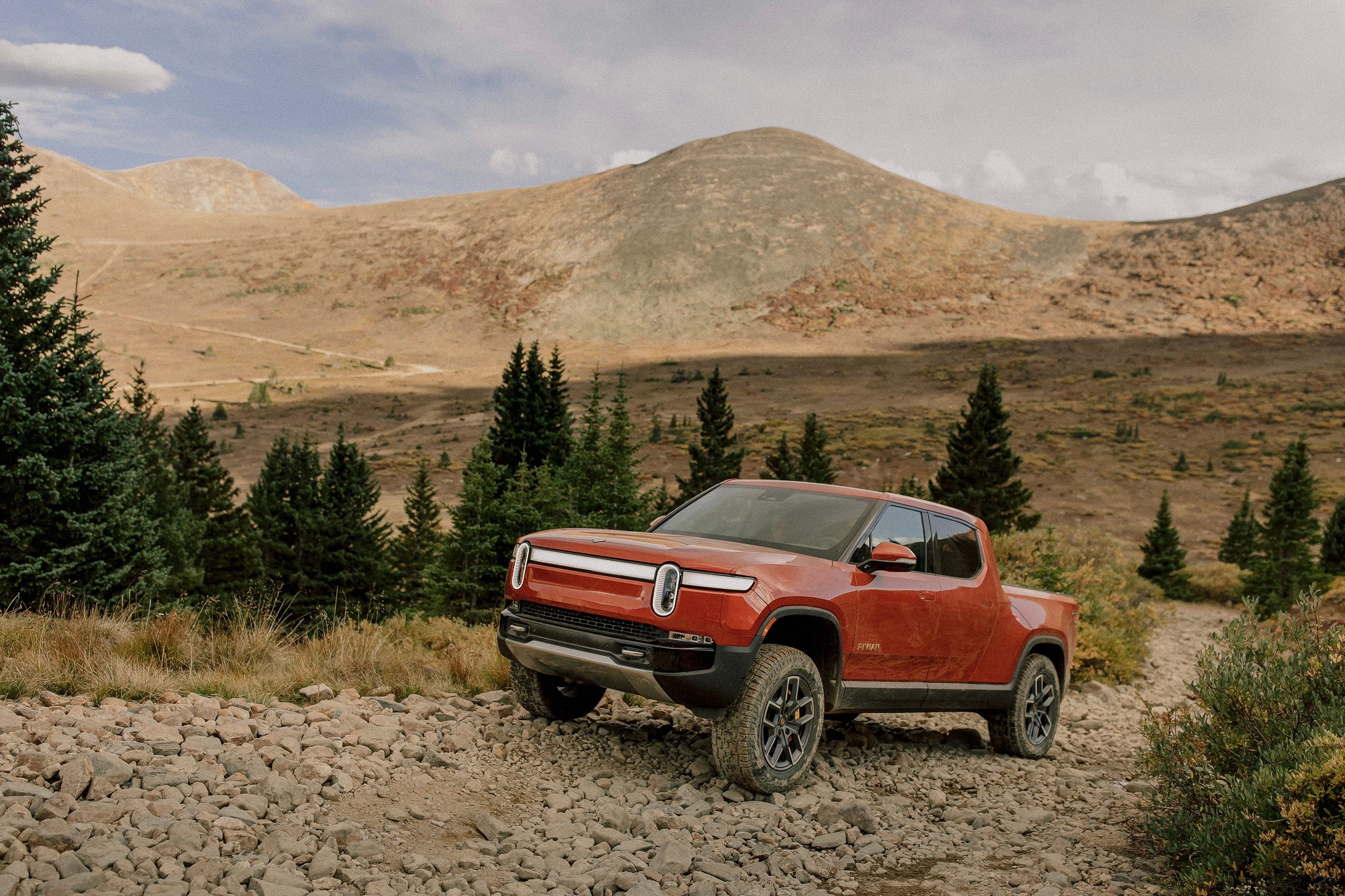 2022 Rivian R1T front end angle driving up a rocky path with mountains and trees in the back.
