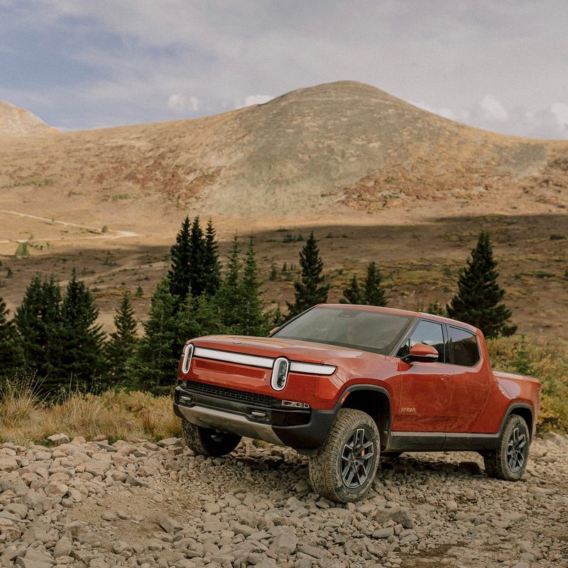 2022 Rivian R1T front end angle driving up a rocky path with mountains and trees in the back.