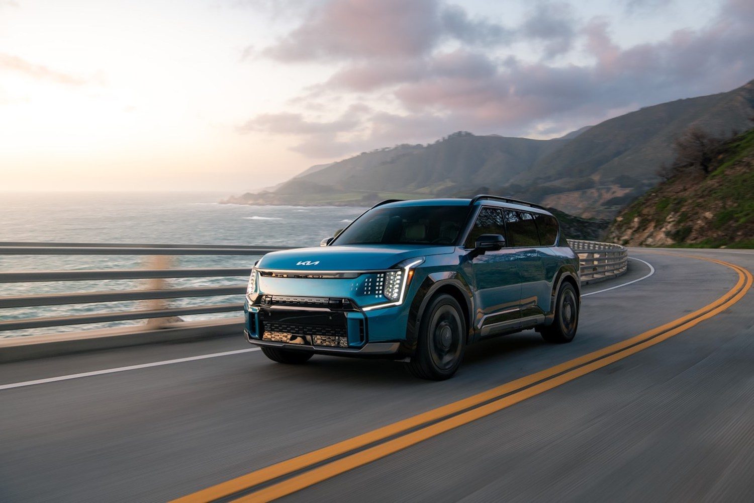2024 Kia EV9 driving down the road with mountains and a body of water in the back.
