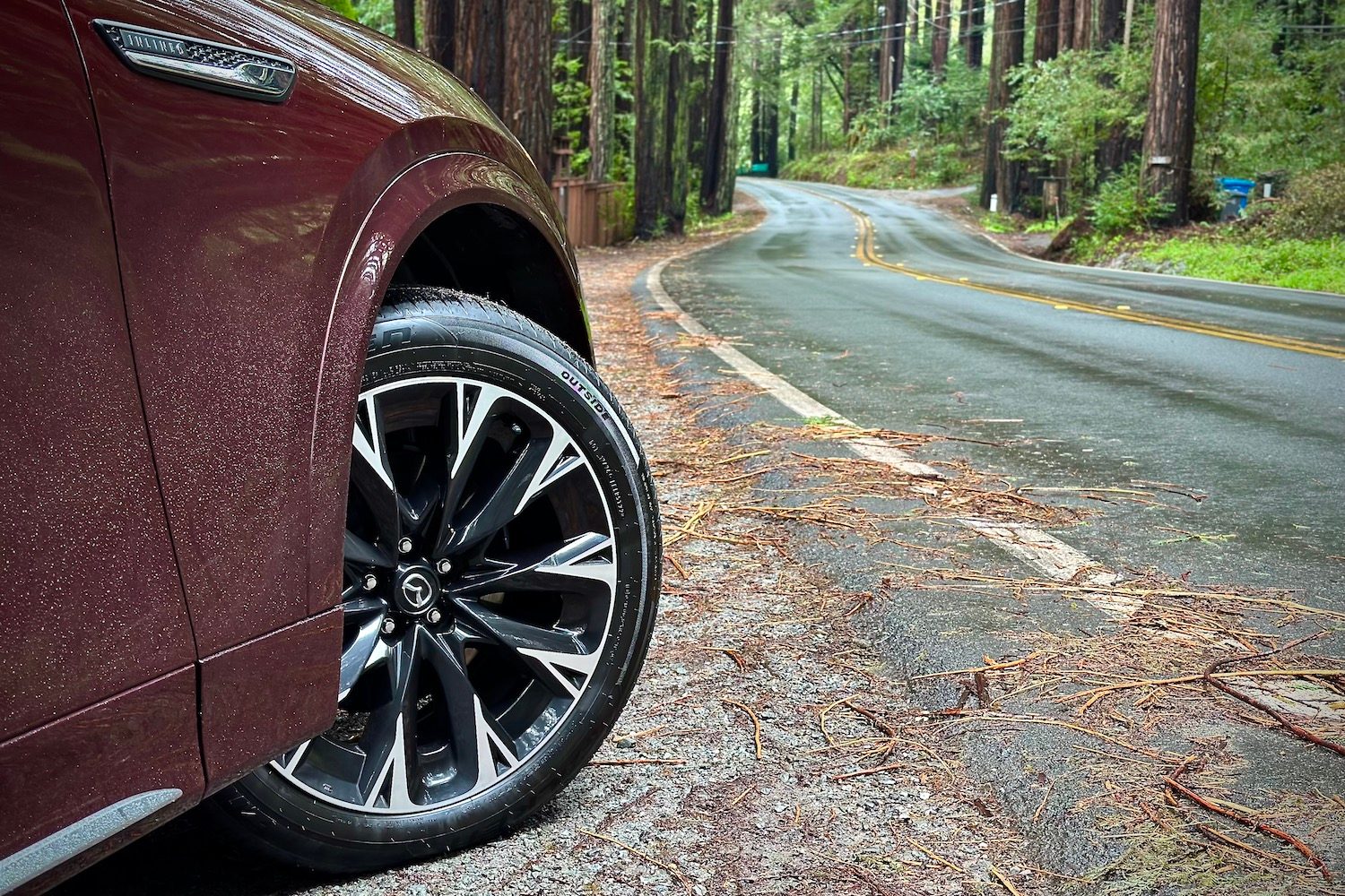 Close up of wheel on the 2024 Mazda CX-90 parked on the side of the road in front of trees.