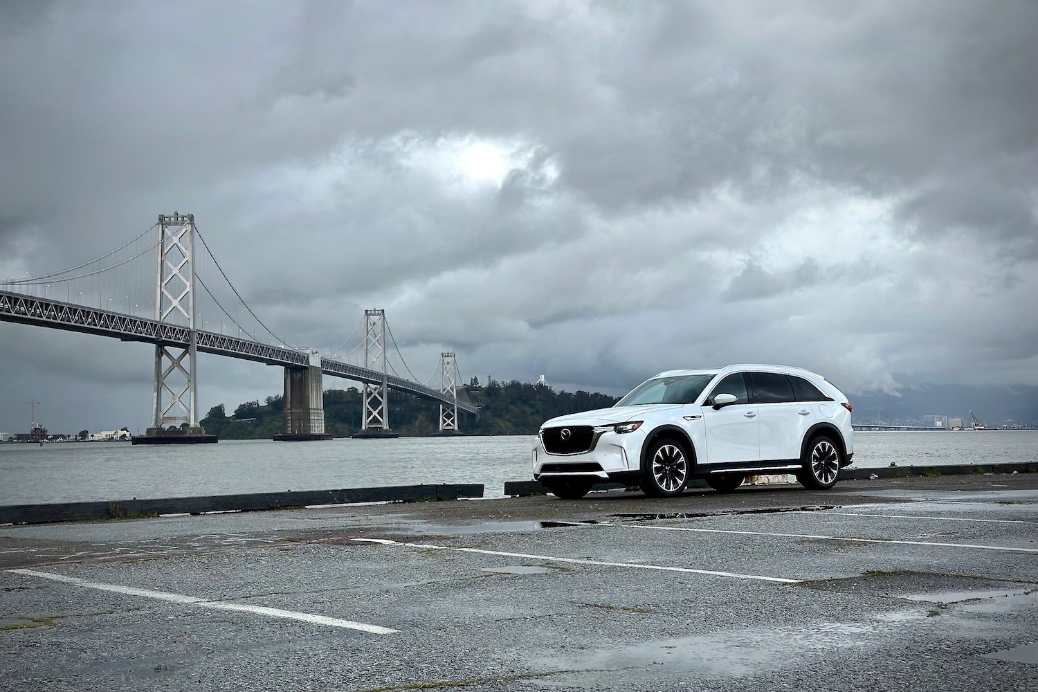 Side profile of the 2024 Mazda CX-90 PHEV parked in front of a bridge with dark clouds.