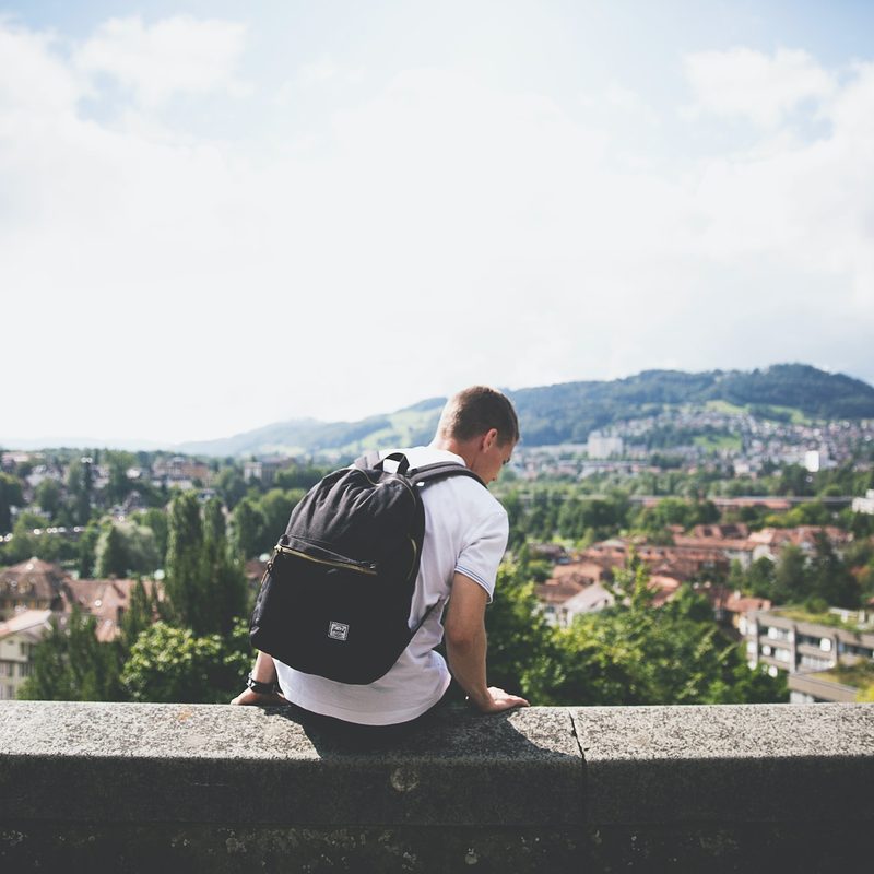 Man with a backpack sitting on a concrete wall overlooking a mountain and urban landscape