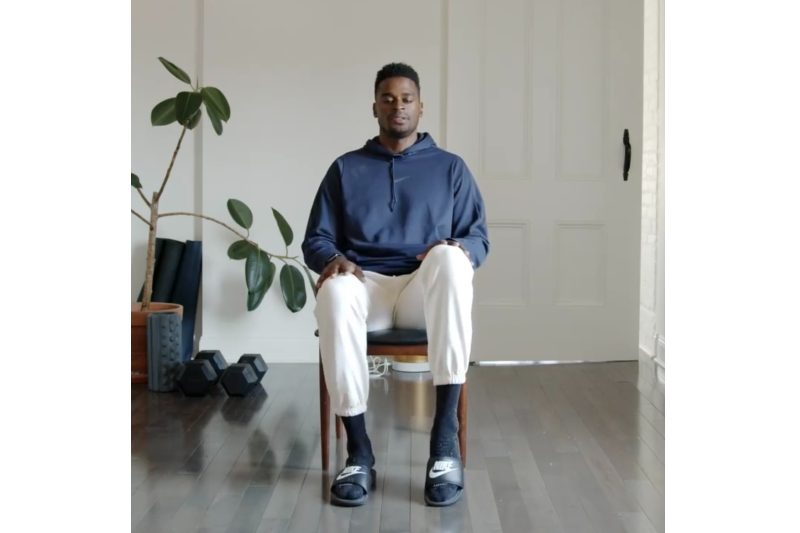 Joe Holder sitting in a chair performing a workout in a living room.
