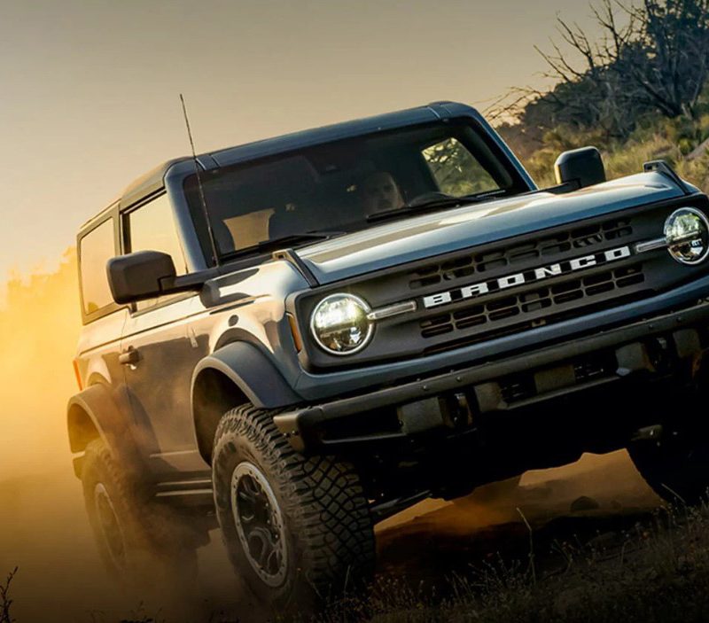 Ford Bronco driving on a dusty backcountry road with sun glinting in the background.