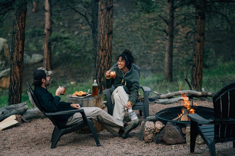 A pair of people enjoy a campfire and a bottle of TINCUP whiskey.