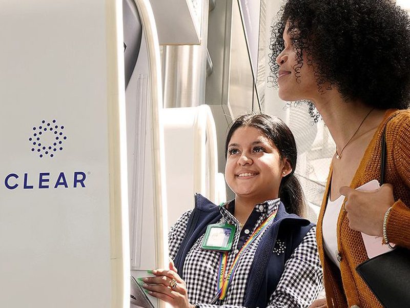 CLEAR program representative helping a traveler at an airport kiosk.