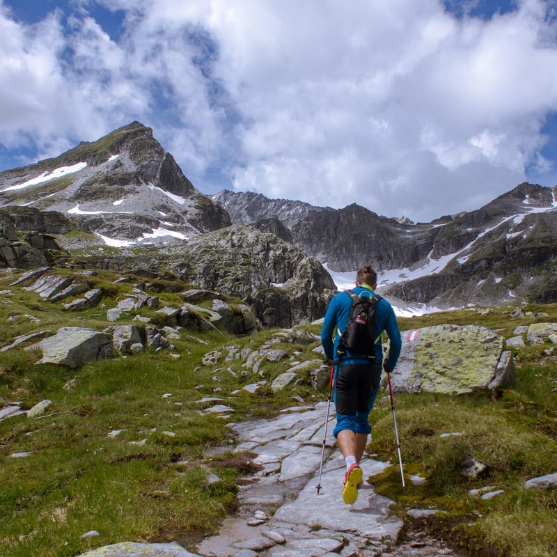 A man hikes up a trail towards a set of snow-topped mountains.
