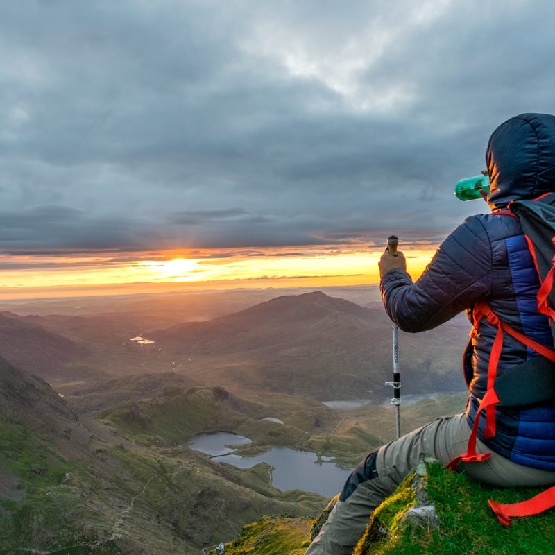 A man sits on a mountain ledge looking out over the sunset.