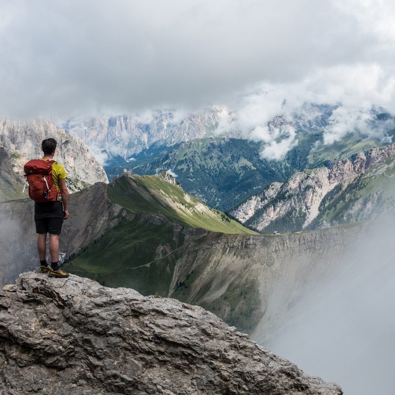 A man with a red backpack stands on a mountaintop looking over more mountains.