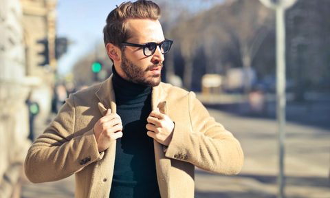 A man outside fixing his coat and wearing glasses.