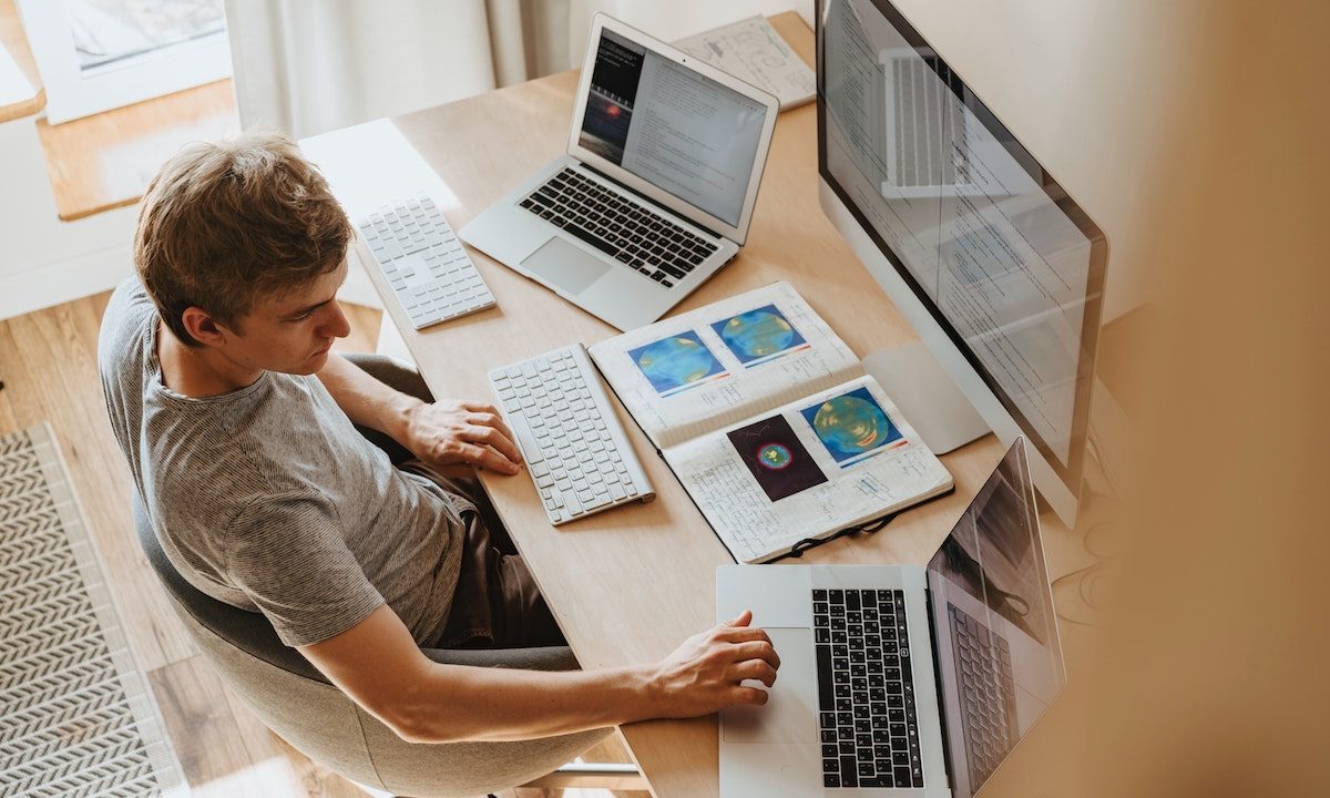 man at wooden desk in front of 3 computer screens