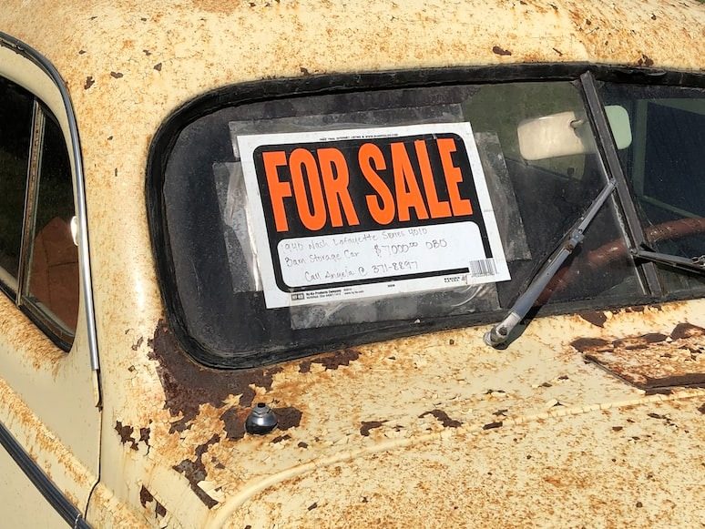 A rusted vintage car with a for sale sign attached to it.