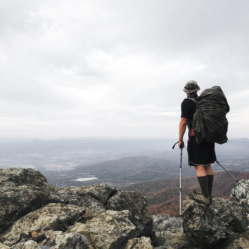 A man stands high up on a trail with hiking poles and a backpack.