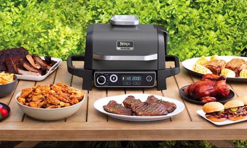 The Ninja Woodfire Outdoor Grill and Smoker on a patio table with plates of food.