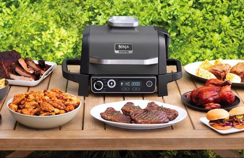 The Ninja Woodfire Outdoor Grill and Smoker on a patio table with plates of food.