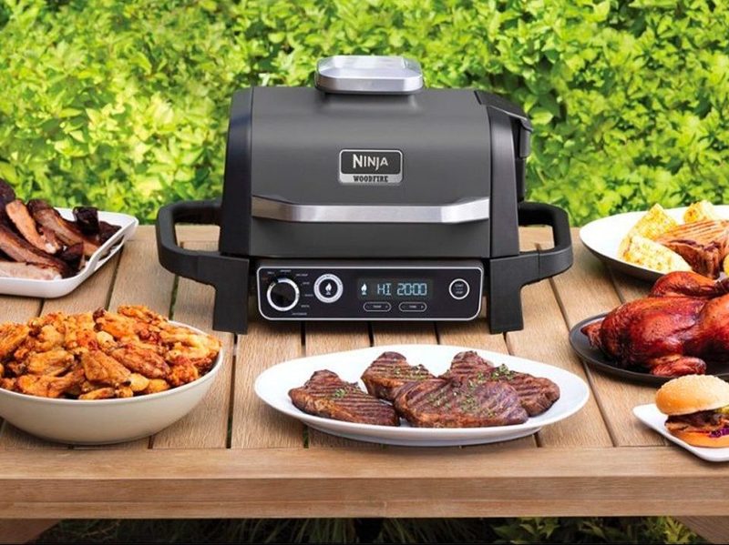 The Ninja Woodfire Outdoor Grill and Smoker on a patio table with plates of food.