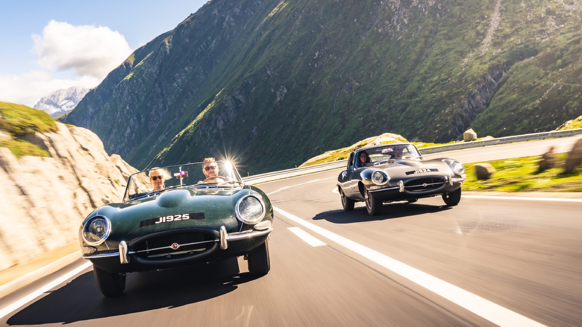 Two Jaguar E-types, a roadster and coupe, driving down a mountain road at speed.