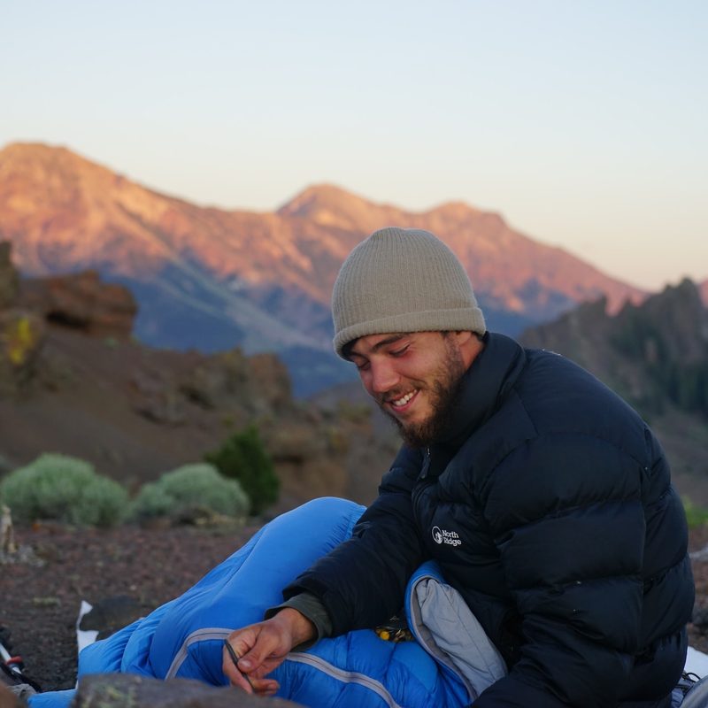 A man sits outside in his sleeping bag overlooking the mountains.