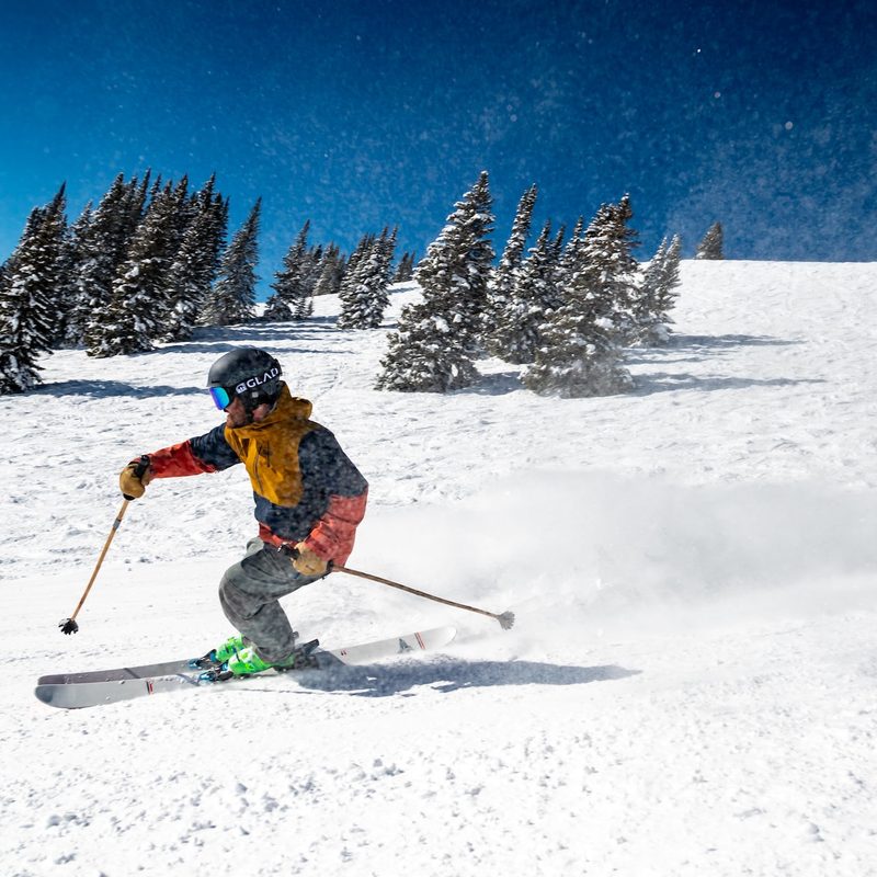 A man skis on a clear run, with mountains and trees behind him.