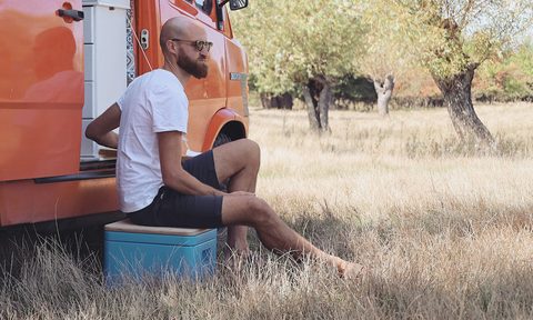 Young bearded man sitting on a Trelino portable composting toilet next to an orange campervan.